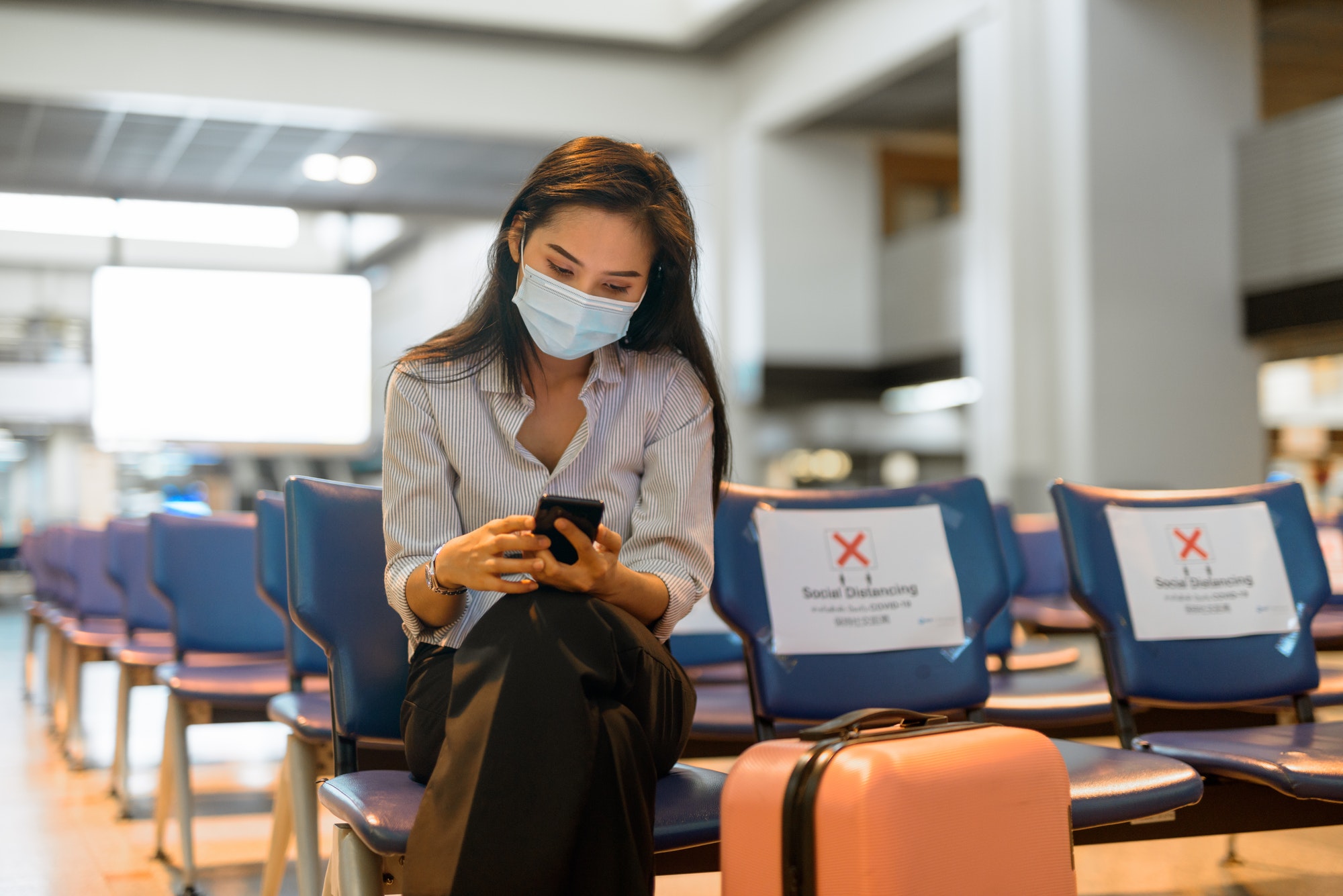 Young Asian tourist woman with mask using phone and sitting with distance at the airport